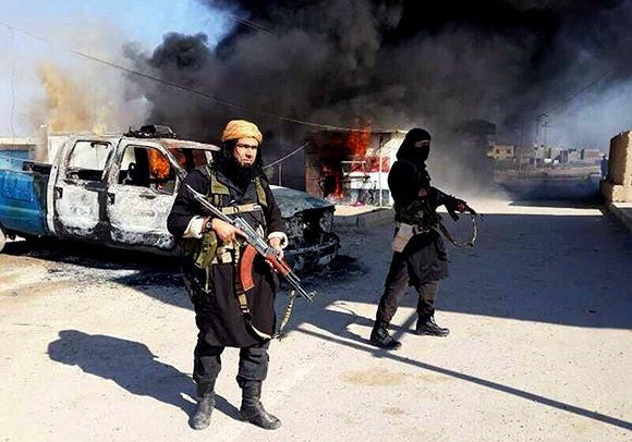 ISIS militants walk armed through a burning street beside a destroyed vehicle.