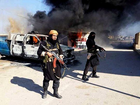 ISIS militants walk armed through a burning street beside a destroyed vehicle.