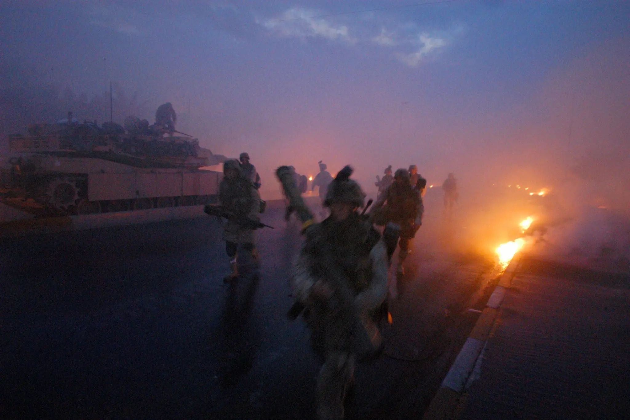 U.S. Marines move through smoke and burning debris during intense urban combat in the Second Battle of Fallujah, Iraq, as an M1 Abrams tank advances behind them at dawn amid the chaos of Operation Phantom Fury.