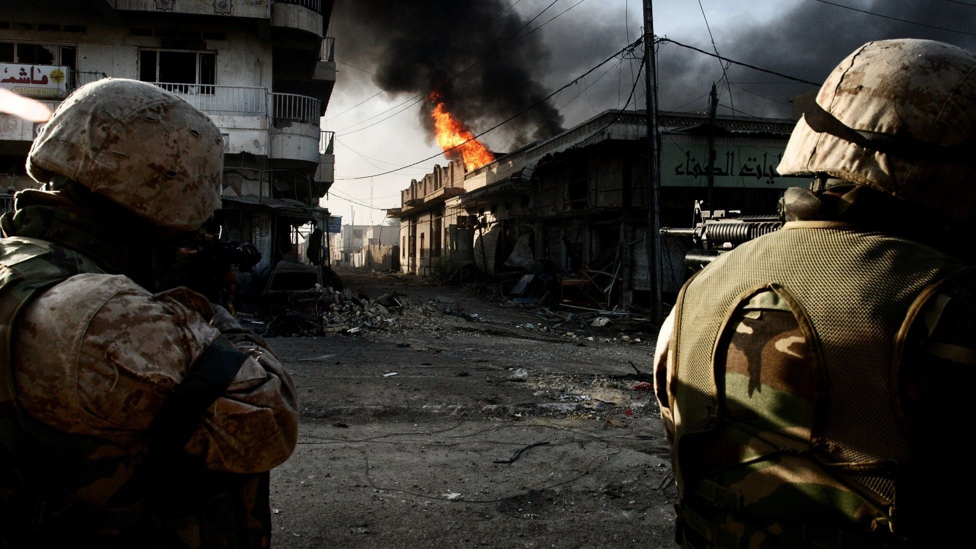 U.S. Marines move through the streets of Fallujah during intense urban combat as flames and black smoke rise from damaged buildings in the background.