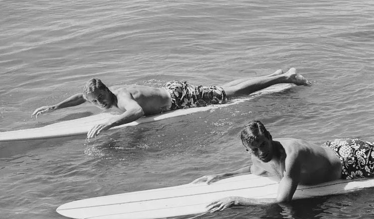 Two friends rest side by side on surfboards in calm water, illustrating themes of friendship, shared experiences, and the side-by-side connection explored in Elliot Ackerman's essay A Man Should Know: How to Be a Friend.