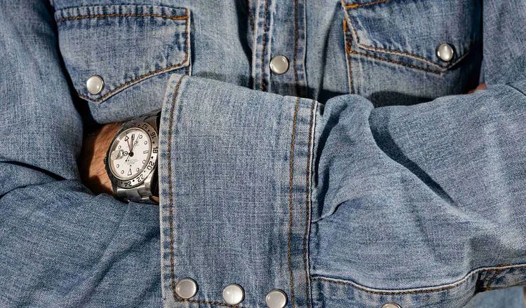 A wristwatch peeks out from beneath the cuff of a denim jacket, illustrating themes of craftsmanship, personal history, and meaningful possessions explored in Elliot Ackerman's essay A Man Should Know: How to Own a Watch.
