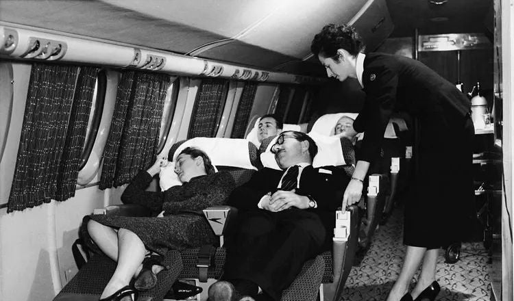 Passengers rest aboard a long-distance aircraft while a flight attendant assists travelers during an overnight journey, illustrating themes of air travel, comfort, and travel etiquette explored in Elliot Ackerman's essay A Man Should Know: How to Travel.