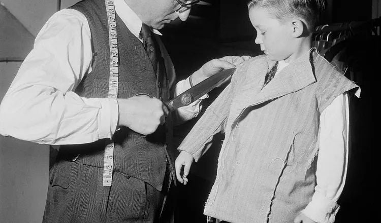 A tailor measures and fits a young boy for a formal jacket, illustrating themes of menswear, tailoring, formal dress, and learning the traditions of dressing well discussed in Elliot Ackerman's essay A Man Should Know: How to Wear a Tuxedo.
