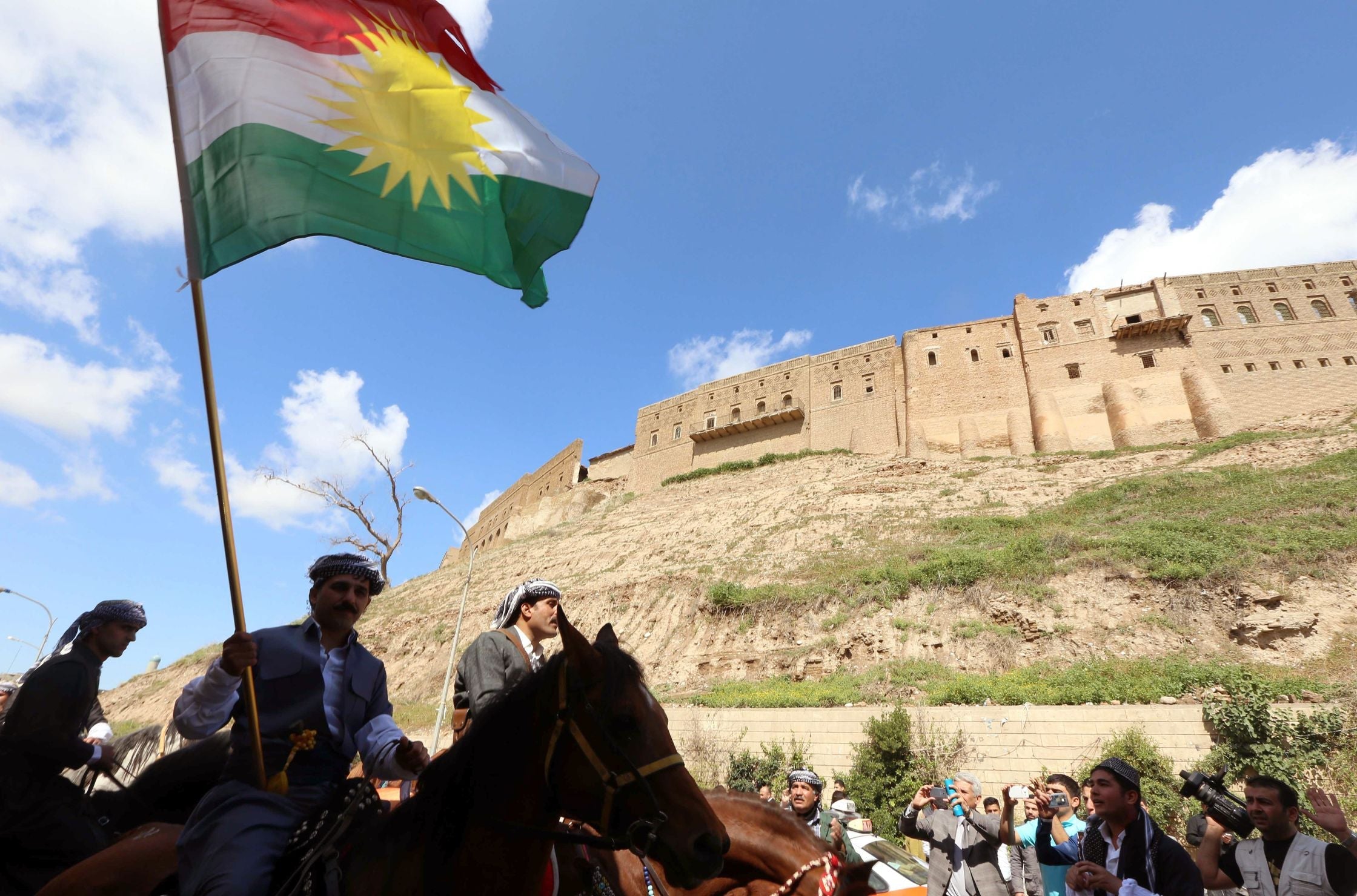 The Kurdish flag flies before the Citadel of Erbil, with men on horseback in traditional dress.