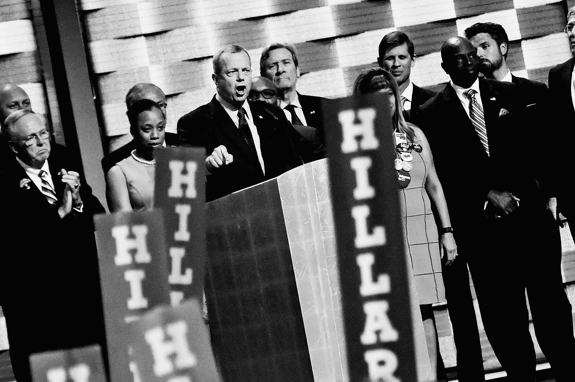 Black-and-white photo of a speaker at a podium surrounded by "Hillary" signs at the 2016 Democratic National Convention.