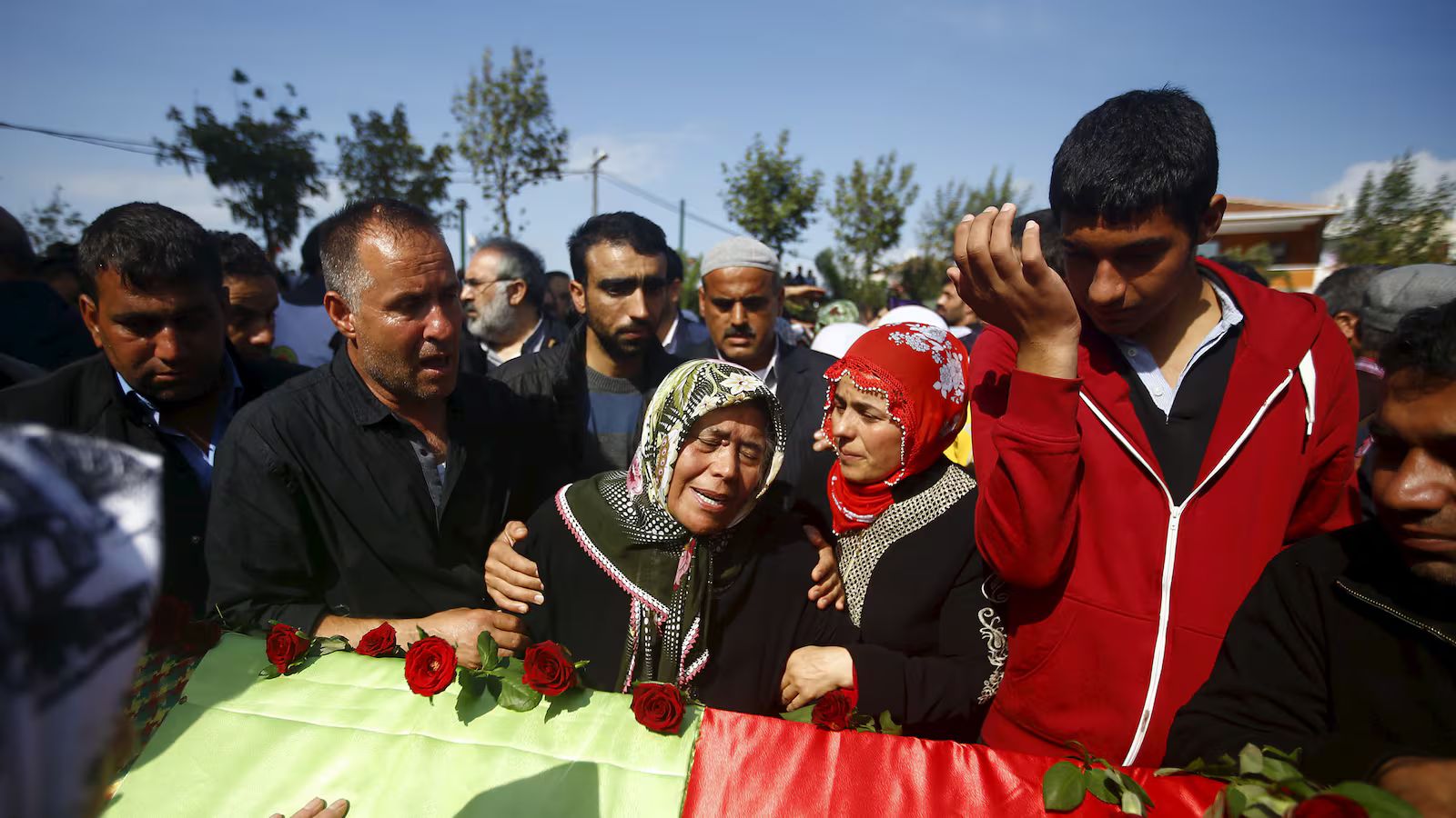 Grieving family members gather around a coffin draped with a green, yellow, and red cloth during a funeral procession following the 2015 Ankara bombing in Turkey.