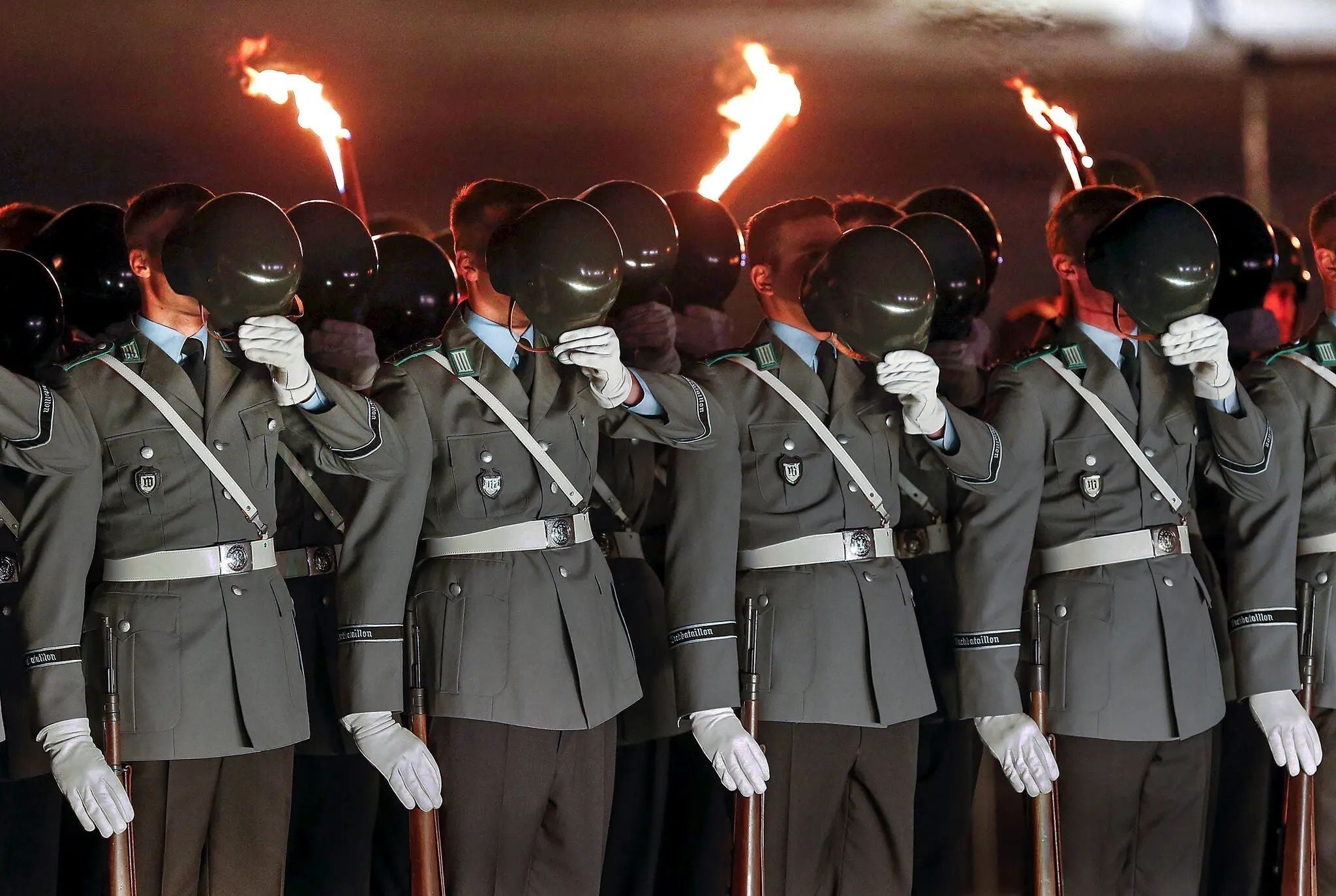 German Bundeswehr soldiers in dress uniform participate in a nighttime military ceremony, holding their helmets before their faces as ceremonial torches burn behind them.