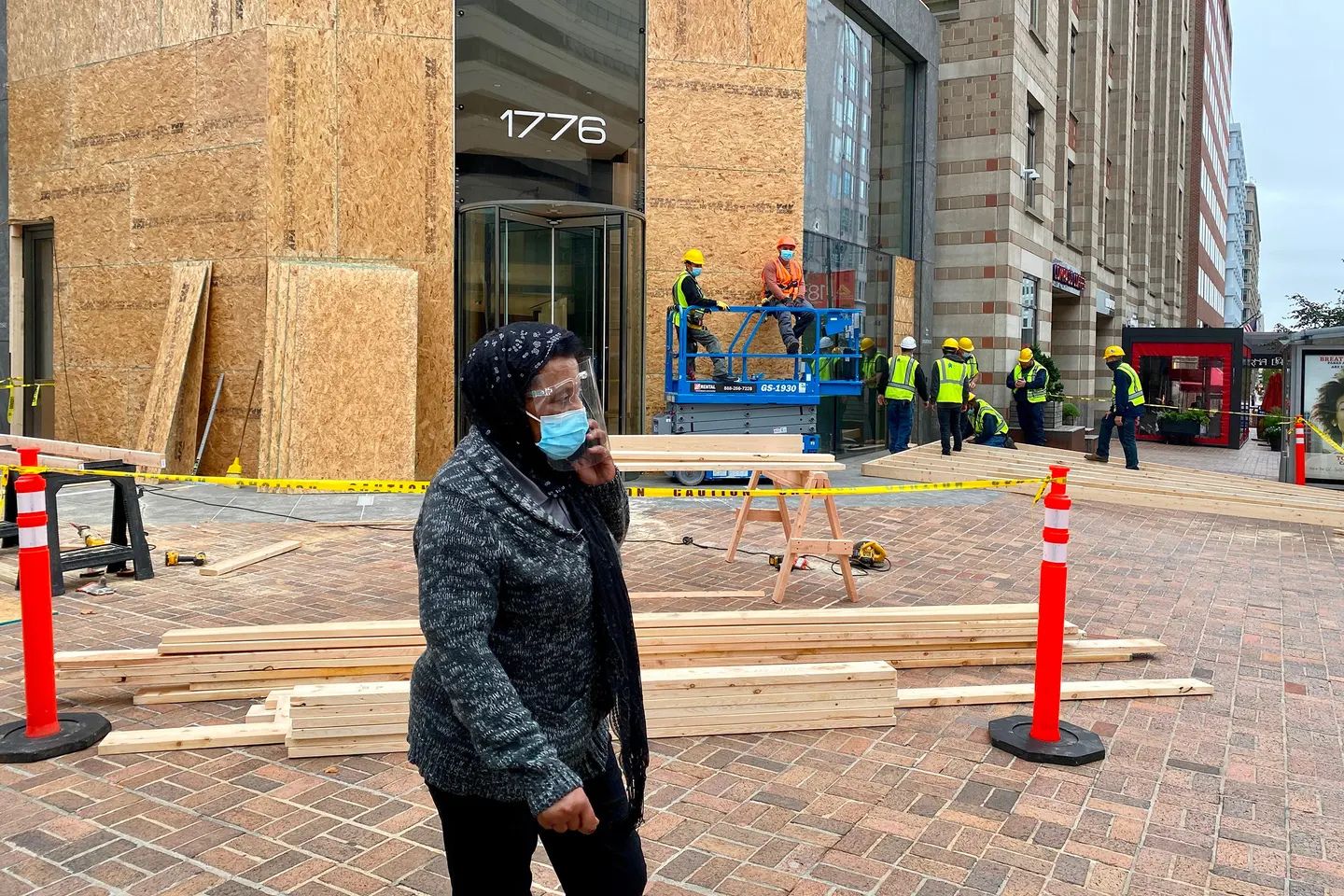 Workers board up the exterior of a downtown office building with plywood while a masked pedestrian walks past, illustrating preparations for potential civil unrest ahead of the 2020 U.S. presidential election.