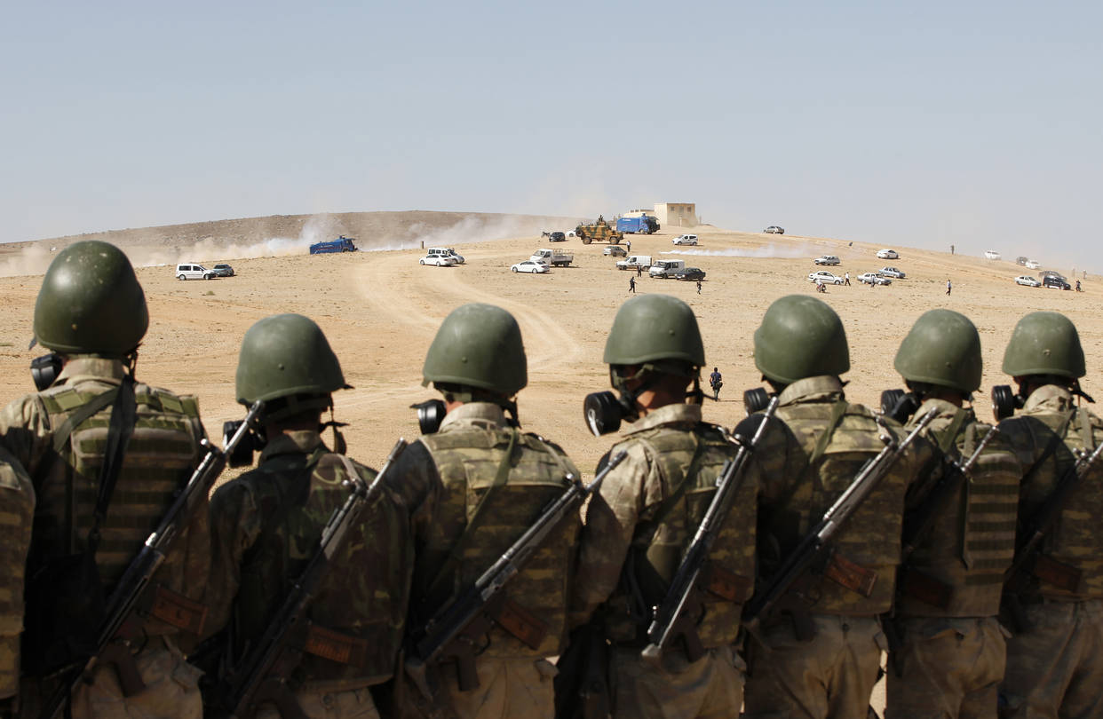 Turkish soldiers stand watch over a desert border checkpoint, with vehicles and smoke visible in the distance.