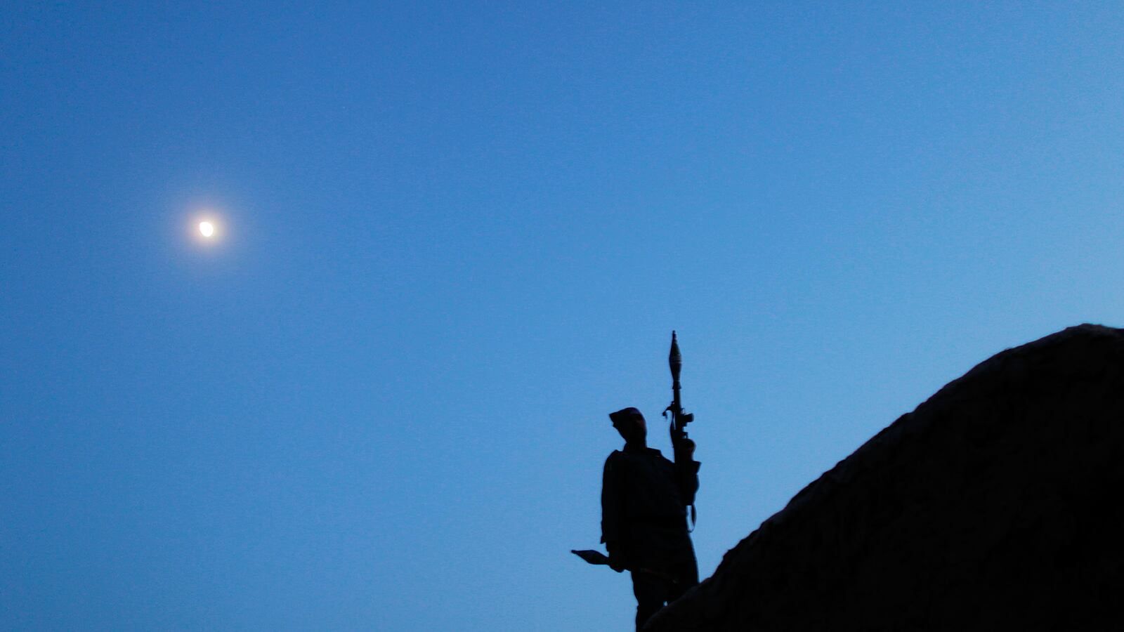 Silhouette of an armed Afghan fighter standing atop a rocky ridgeline at dusk beneath a bright moon, holding an RPG launcher against a deep blue twilight sky.