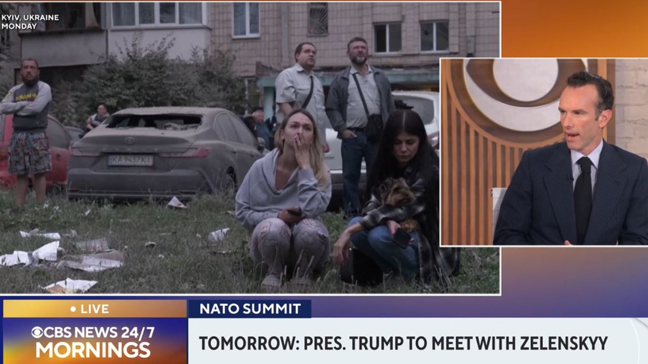 Split-screen from a CBS News 24/7 broadcast showing civilians gathered outside damaged apartment buildings in Kyiv, Ukraine, following a Russian attack, alongside CBS News contributor Elliot Ackerman.