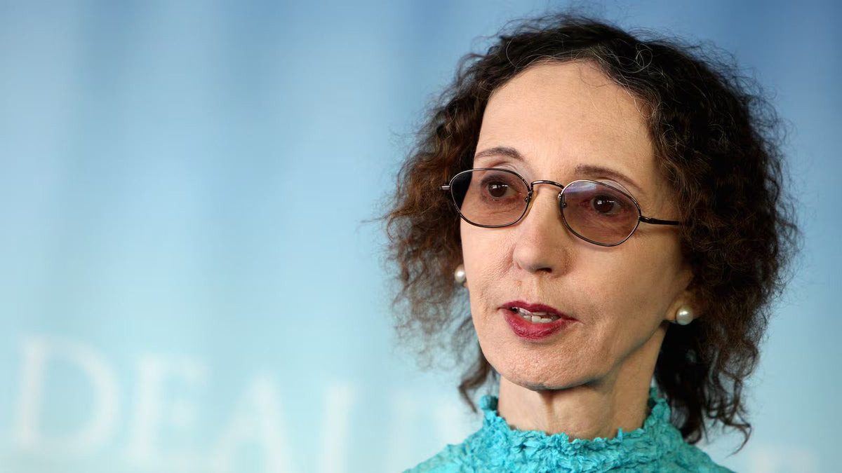 Portrait of acclaimed American novelist Joyce Carol Oates wearing round glasses, pearl earrings, and a turquoise blouse, speaking during an interview against a softly blurred blue background.
