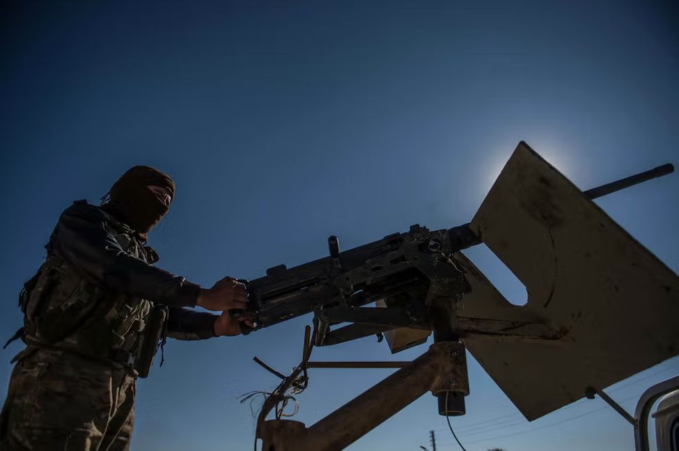 Masked Islamic State (ISIS/Daesh) fighter operating a mounted heavy machine gun in Syria during the Syrian Civil War, silhouetted against a bright blue sky.