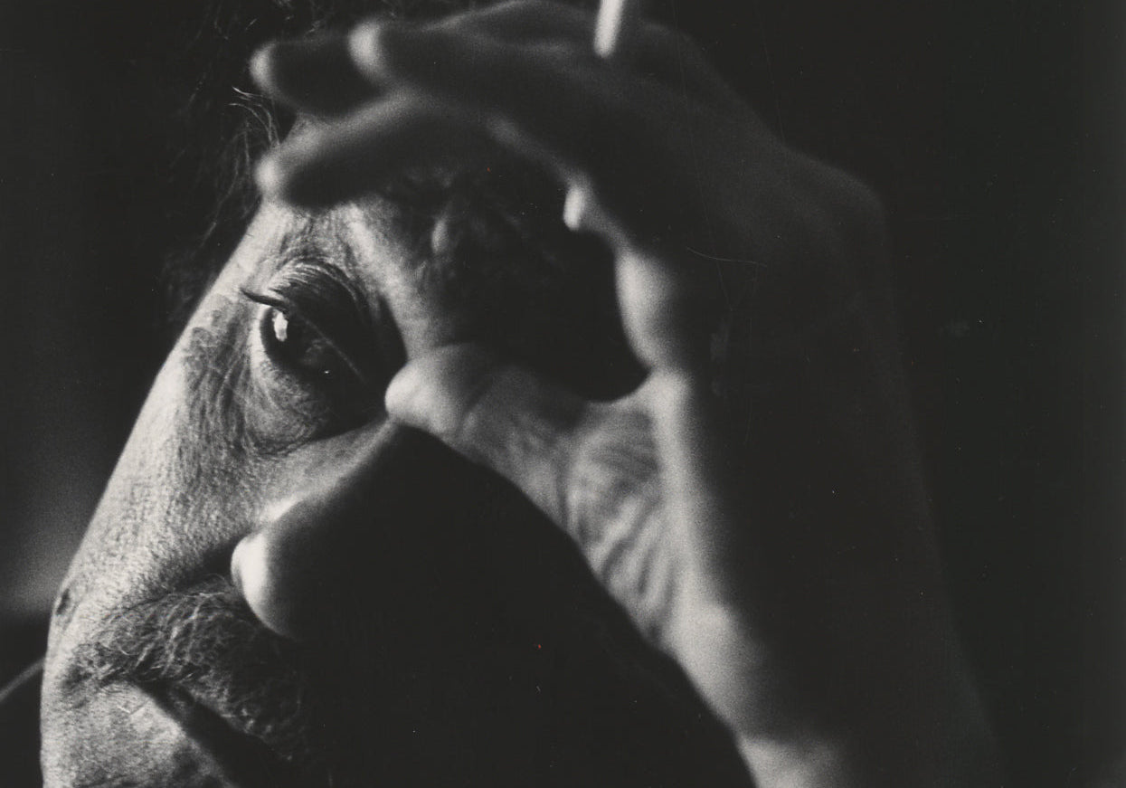 A dramatic black-and-white close-up portrait of a man's face partially obscured by his hand, holding a cigarette.