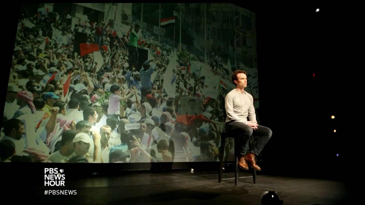 Elliot Ackerman sits on stage during a PBS NewsHour essay with a large image of Arab Spring protesters projected behind him, reflecting on war, revolution, and friendship in Syria and the broader Middle East.