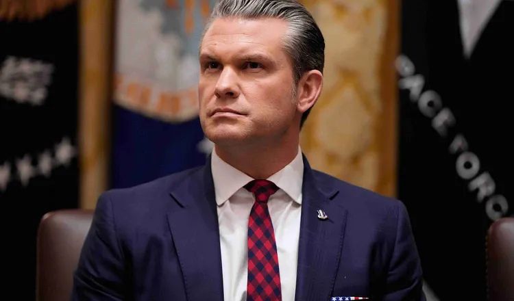 Pete Hegseth, U.S. Secretary of Defense, sits at a conference table in a dark suit and red checkered tie during an official government meeting, with military and departmental flags visible in the background.