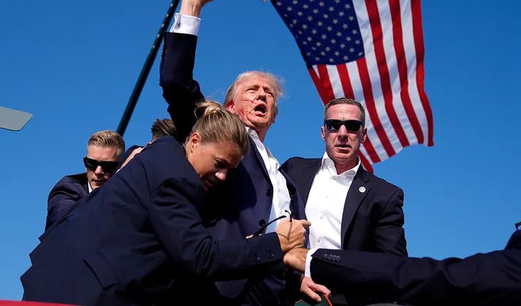 Donald Trump raises his fist and speaks to supporters as U.S. Secret Service agents surround and protect him on stage, with an American flag visible in the background following an assassination attempt at a campaign rally.