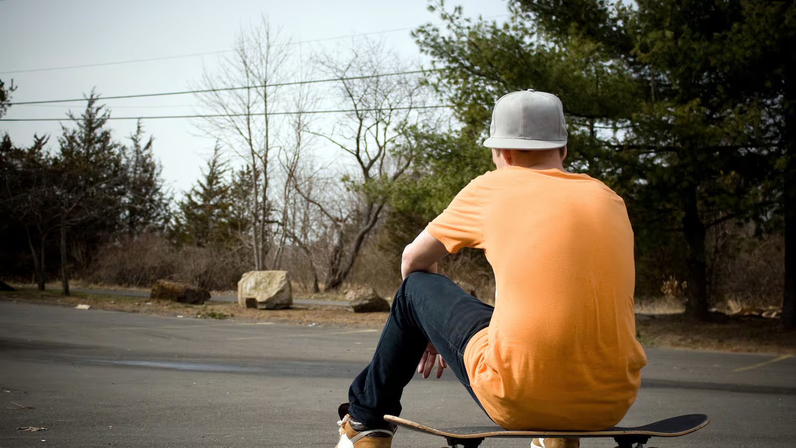 A teenage boy wearing an orange T-shirt, jeans, and a gray baseball cap sits alone on a skateboard in an empty suburban parking lot, viewed from behind.