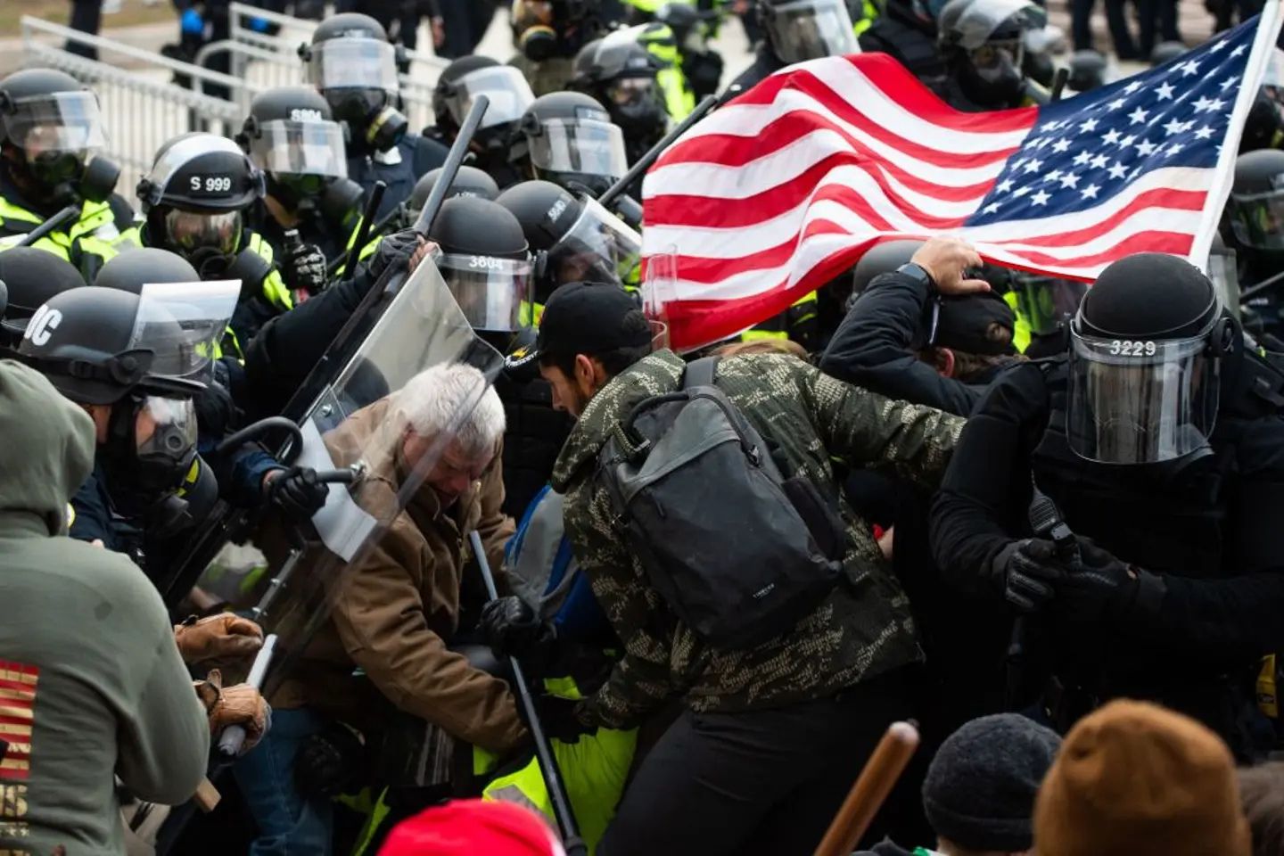 Police in riot gear clash with demonstrators outside the U.S. Capitol during the January 6, 2021 attack, as an American flag waves above the crowd amid shields, helmets, and pushing between law enforcement and protesters.
