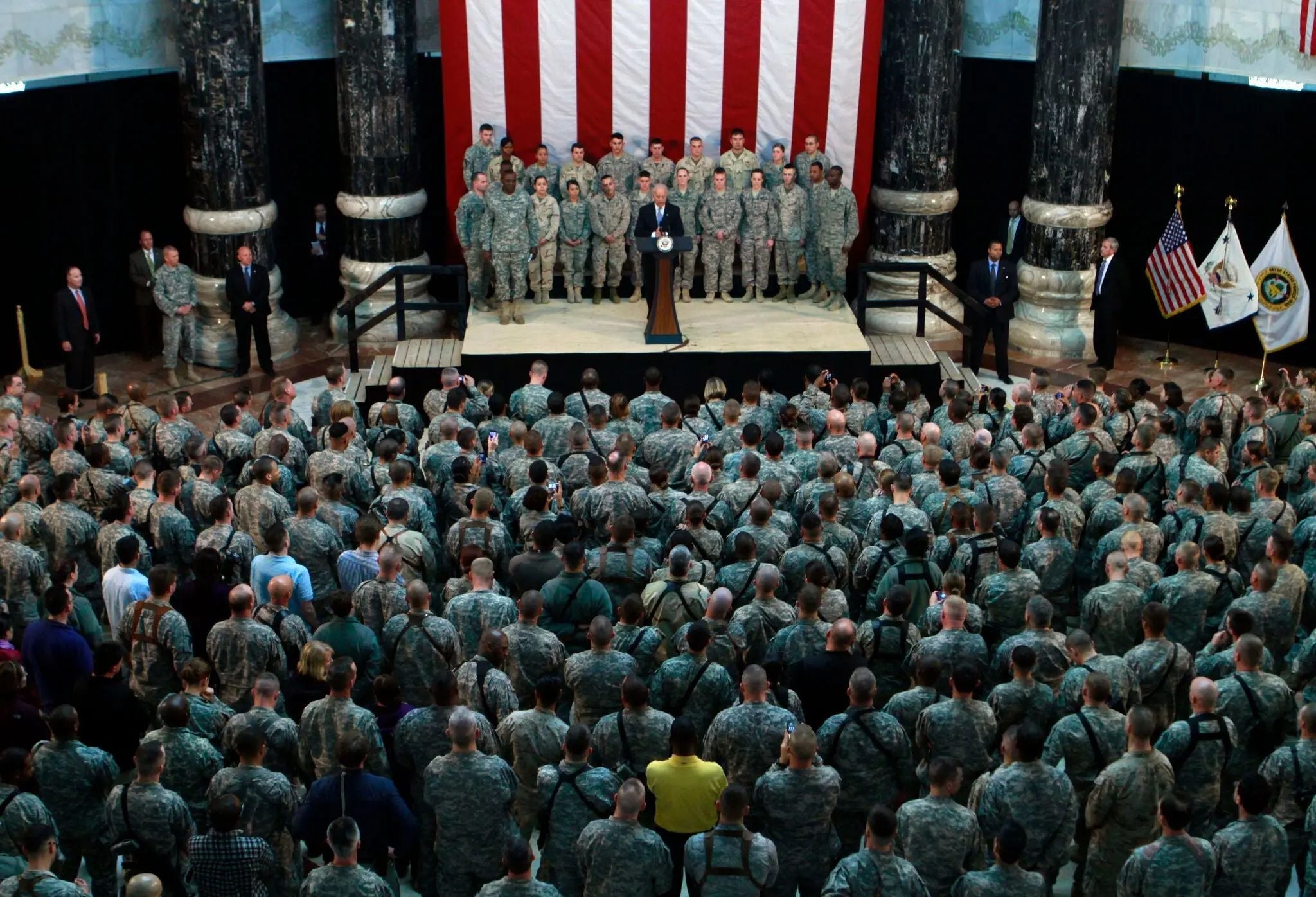 President Joe Biden addresses a large audience of U.S. Army soldiers gathered beneath a giant American flag during a military event, with service members standing behind the podium.