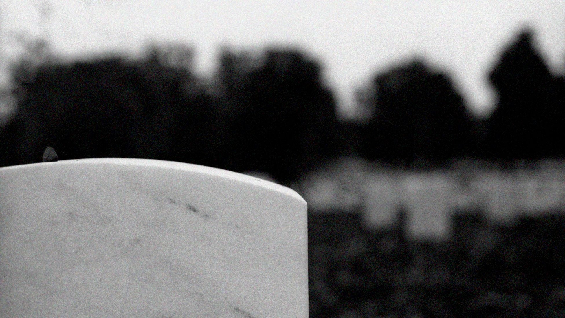 A white marble military headstone stands in a quiet cemetery, with rows of graves blurred in the background.