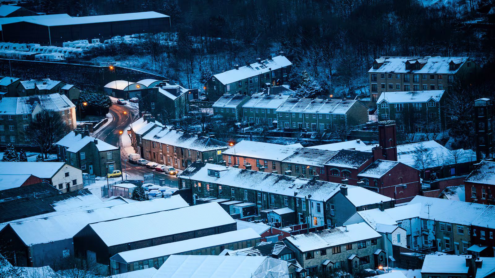 Aerial view of a snow-covered European town at dusk, with rows of terraced houses, winding streets, and warm streetlights glowing against the cold blue landscape.
