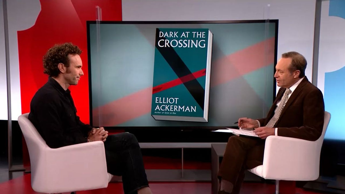 Elliot Ackerman speaks with PBS NewsHour correspondent Jeffrey Brown about his novel Dark at the Crossing, with the book cover displayed behind them during a discussion of the Syrian civil war, refugees, and the power of fiction to humanize conflict.