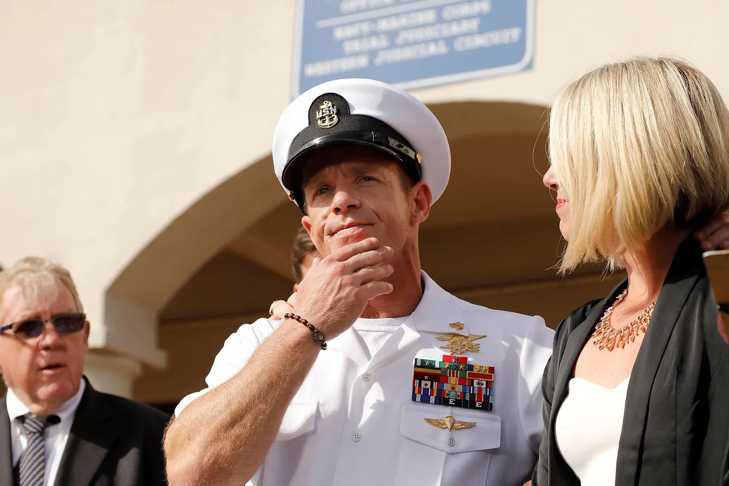 Navy SEAL Chief Edward Gallagher stands in dress white uniform alongside his wife after legal proceedings, wearing his military decorations and Trident insignia.