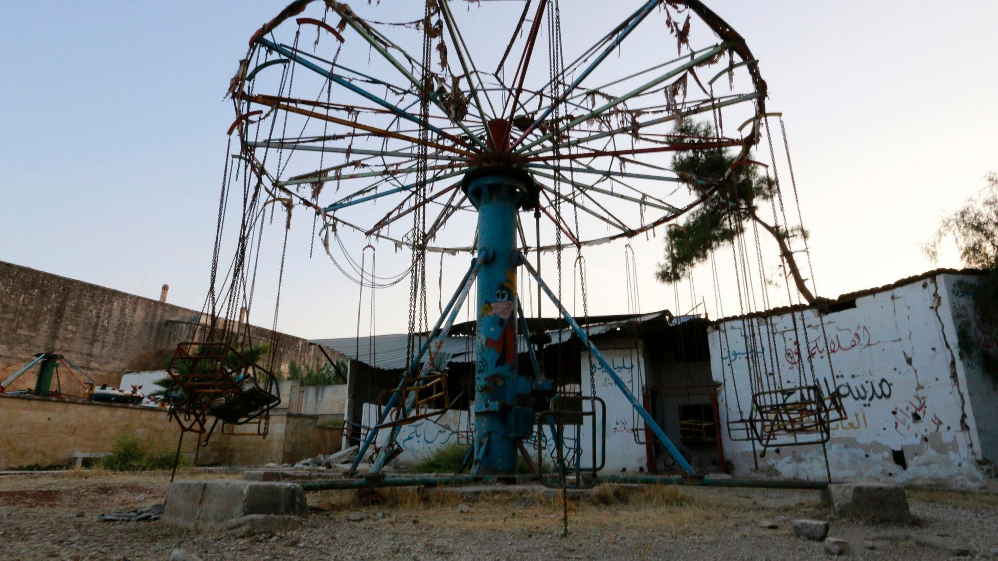 Abandoned amusement park swing ride in a war-damaged Syrian city, with empty chain-hung seats, faded paint, and graffiti-covered walls, symbolizing the impact of conflict on civilian life and childhood spaces.