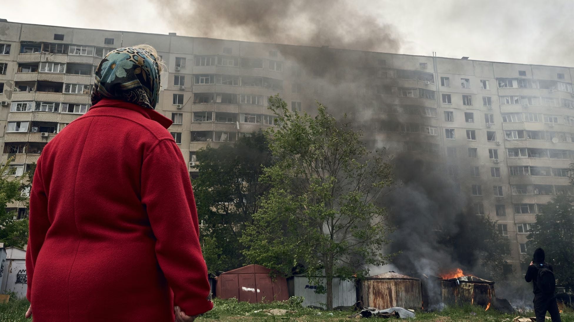 Civilian in a red coat stands before a damaged apartment building in Ukraine as thick black smoke rises from fires caused by a Russian missile strike, illustrating the destruction of urban areas during the ongoing war.
