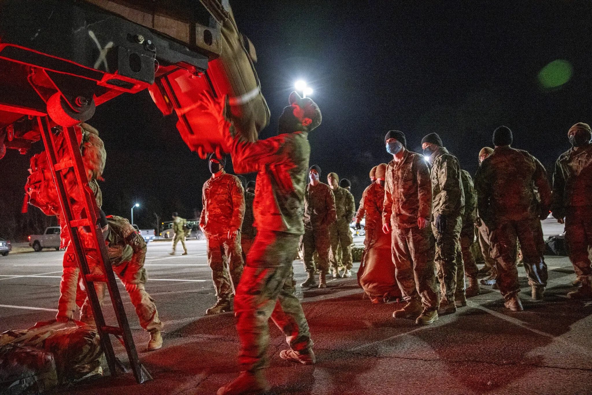 U.S. Army soldiers prepare to board a military aircraft during a nighttime deployment from an airfield, illuminated by red aircraft lighting and floodlights.