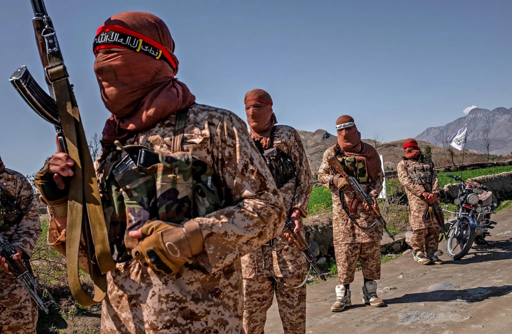 Armed Taliban fighters wearing desert camouflage uniforms and face coverings stand in a rural Afghan landscape with mountains in the background.