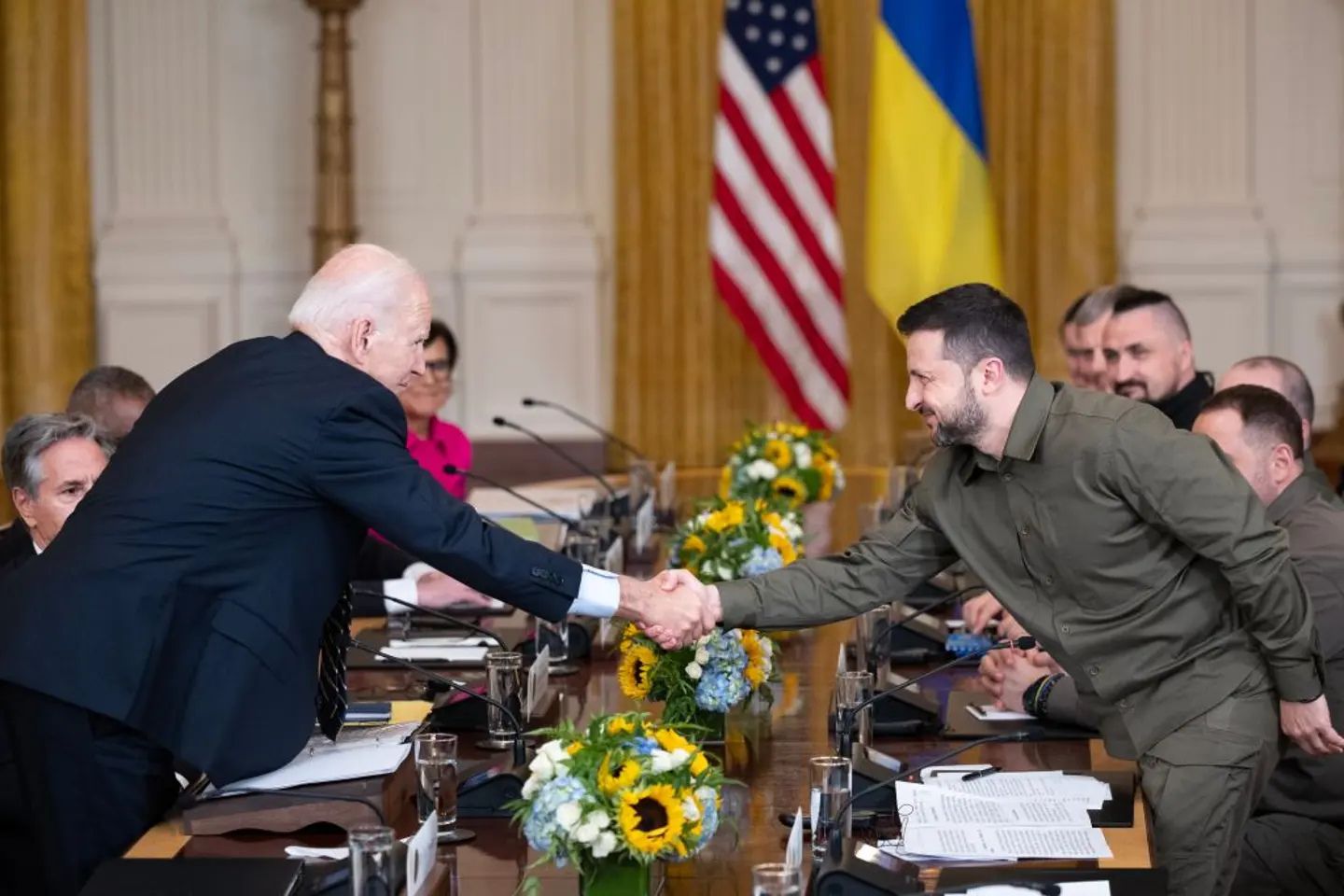 U.S. President Joe Biden shakes hands with Ukrainian President Volodymyr Zelensky during a bilateral meeting, with U.S. and Ukrainian officials seated at a conference table beneath the American and Ukrainian flags.