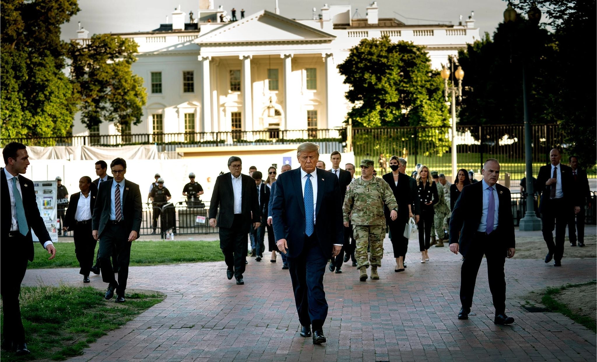President Donald Trump walks with senior administration officials, military personnel, and Secret Service agents outside the White House following unrest in Washington, D.C., with the White House prominently visible in the background.