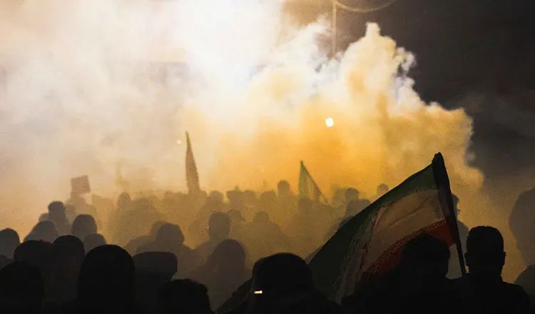 Crowds gather amid smoke and Iranian flags during a nighttime protest in Iran, illustrating themes of regime change, political unrest, and instability discussed in Elliot Ackerman's essay The Case Against the War.