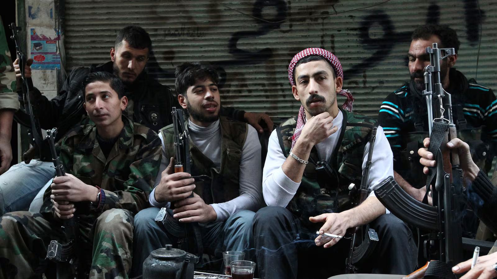 Armed Syrian opposition fighters gather inside a storefront in northern Syria, seated together with AK-pattern rifles while drinking tea and smoking cigarettes.