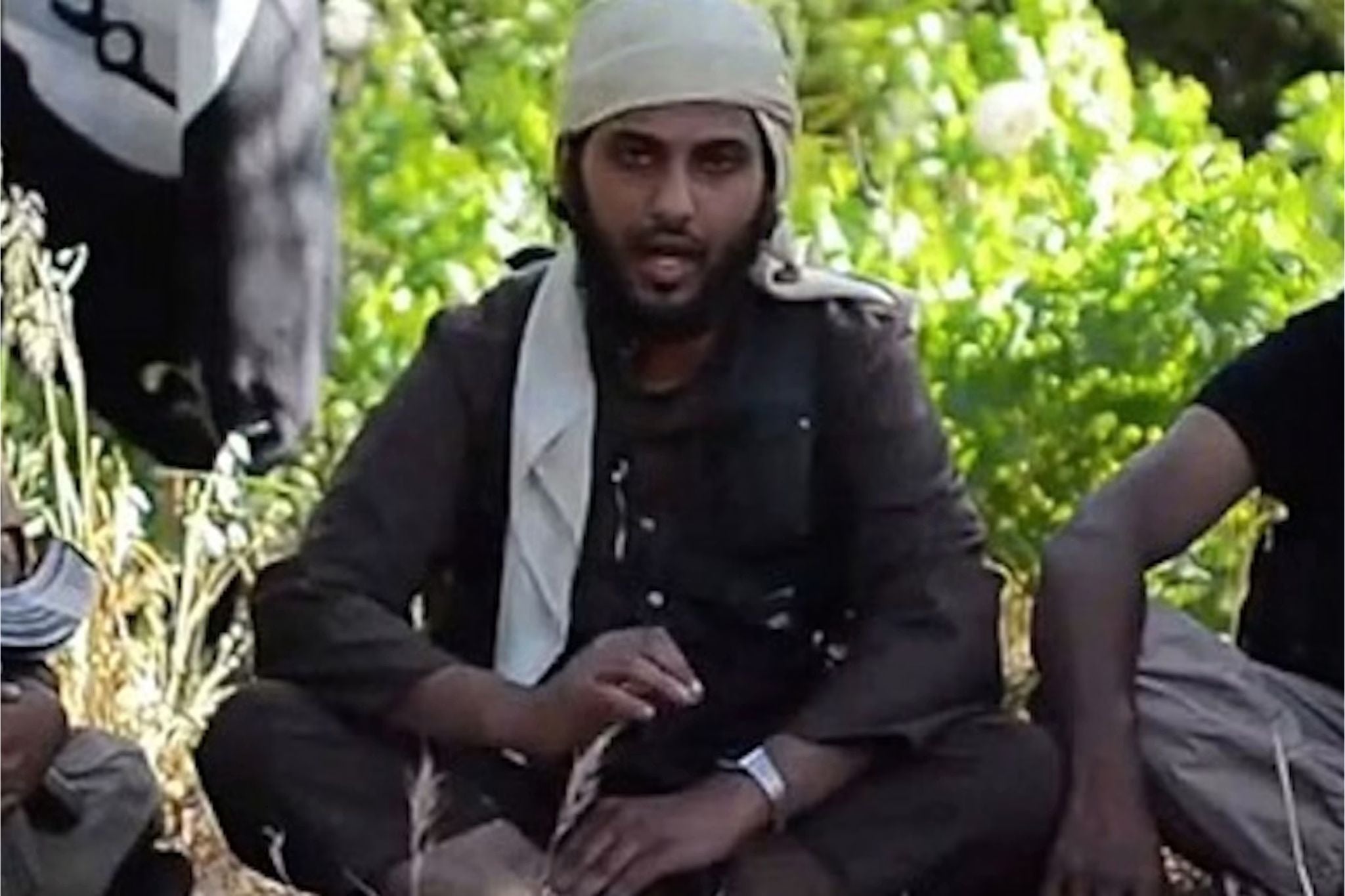 Abu Bakr al-Baghdadi, leader of the Islamic State (ISIS), appears in a propaganda video seated outdoors wearing traditional clothing and a head covering, speaking to the camera with an ISIS flag partially visible beside him.