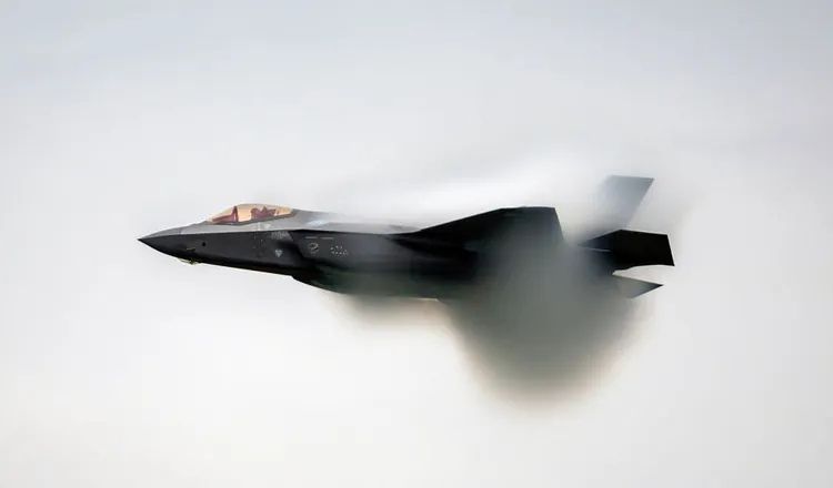 F-35 Lightning II stealth fighter jet flying at high speed, creating a visible vapor cone as it approaches transonic flight during an aerial demonstration.