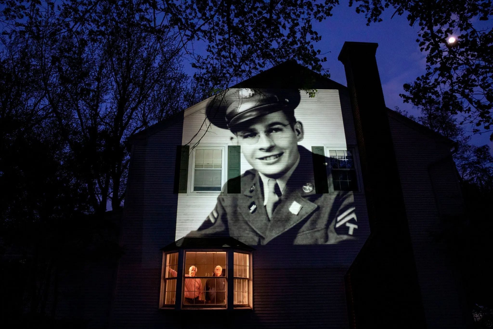 A large black-and-white portrait of a U.S. Army veteran is projected onto the exterior of a suburban home at night, while two family members stand illuminated in a bay window below.