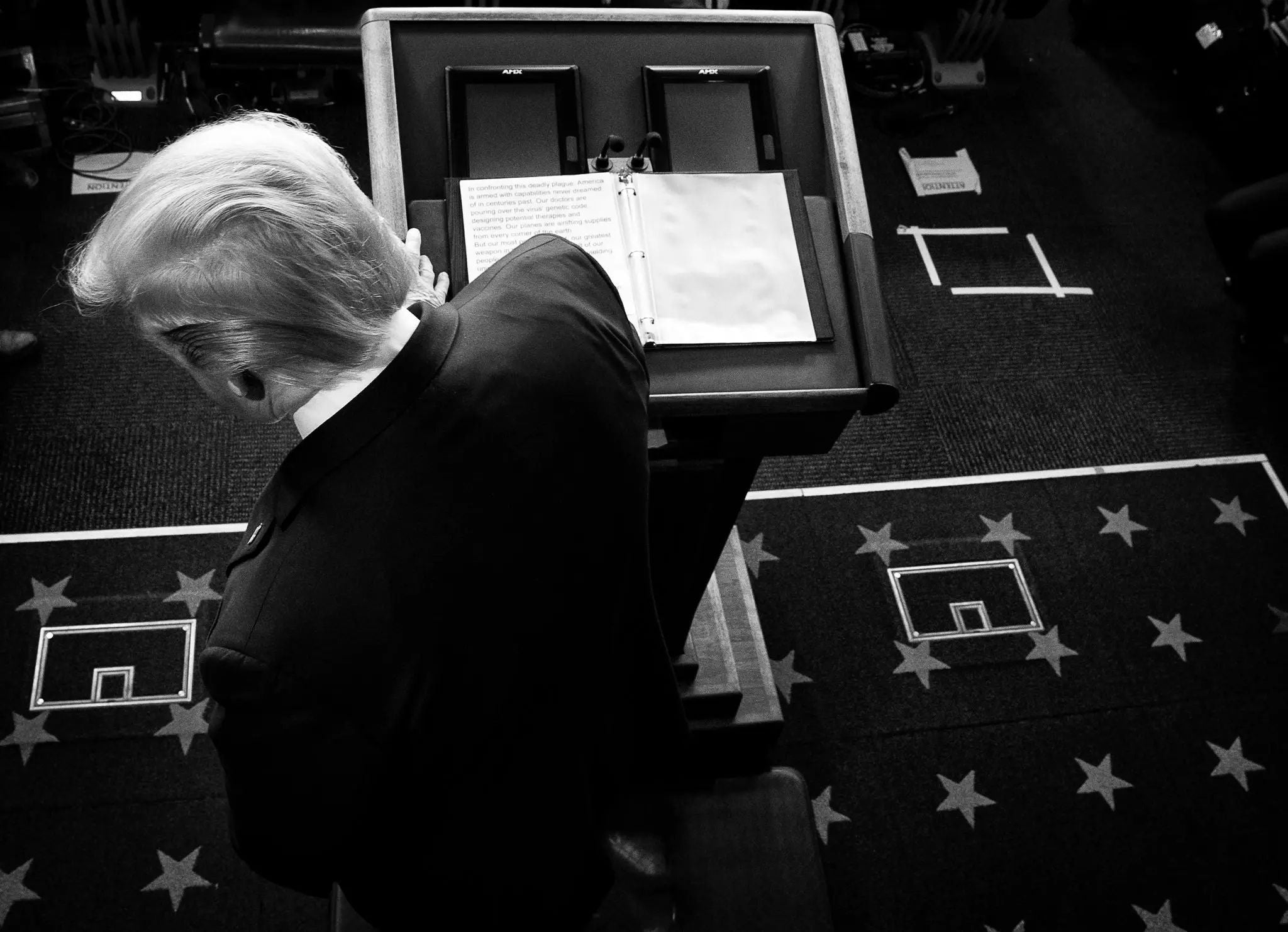 President Donald Trump stands at a White House podium during a COVID-19 press briefing, viewed from above in black and white, symbolizing presidential leadership, crisis communication, and the national response to the coronavirus pandemic.