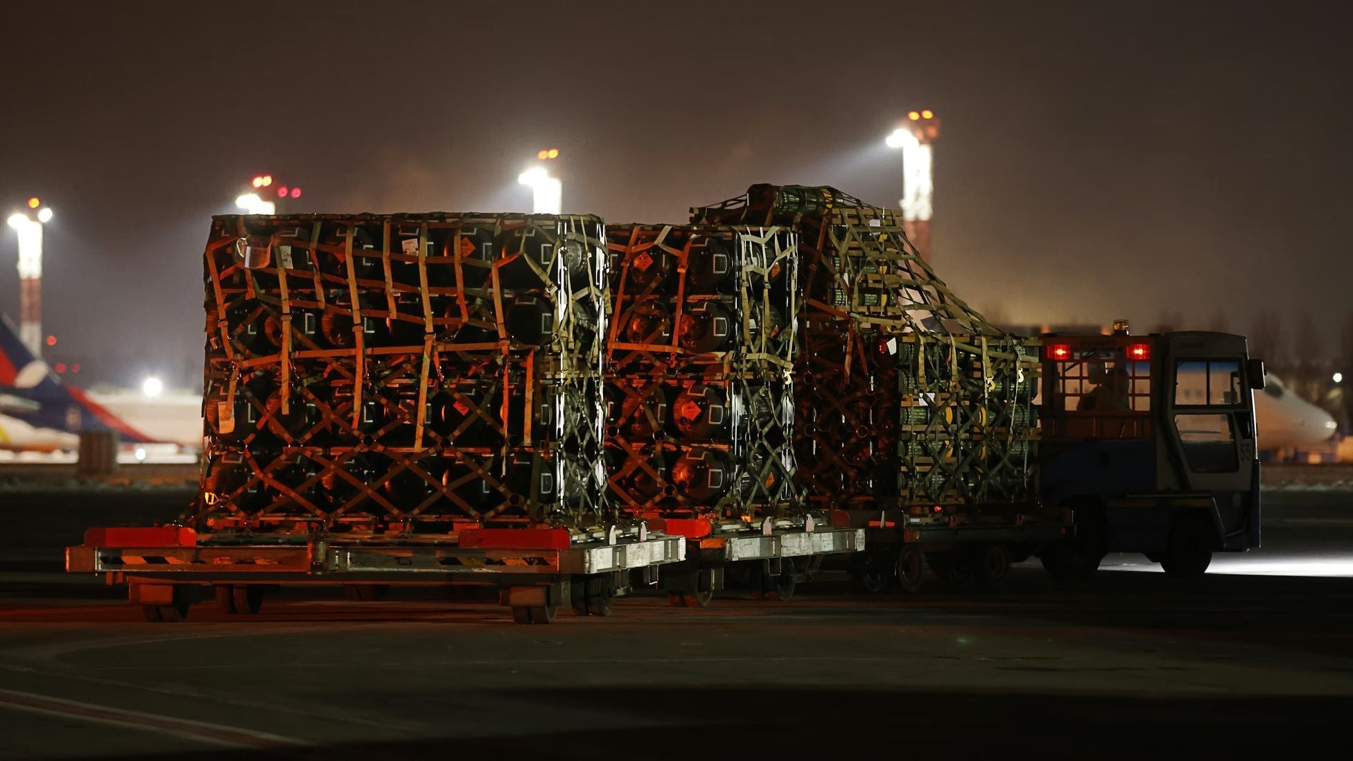 Palletized military cargo secured with netting is transported across an airport tarmac at night, illuminated by floodlights as aircraft and logistics operations continue in the background.