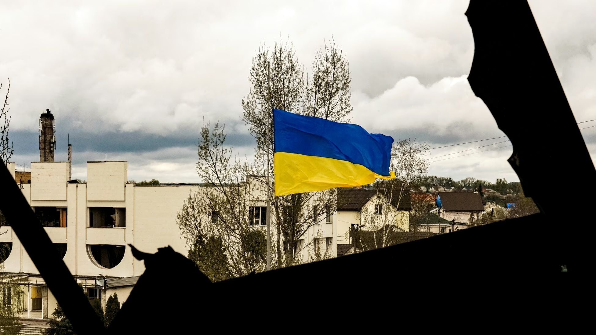 Ukrainian national flag flying above a town damaged by war, viewed through the dark silhouette of a ruined building, symbolizing Ukraine’s resilience and determination amid the ongoing conflict with Russia.