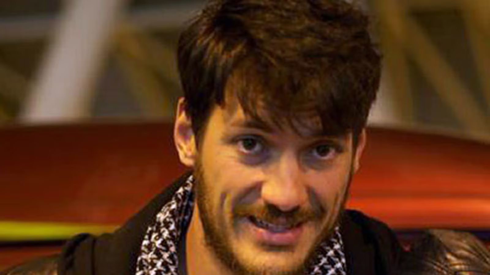 Close-up portrait of American journalist and former Marine Austin Tice wearing a dark jacket and patterned scarf while seated indoors.