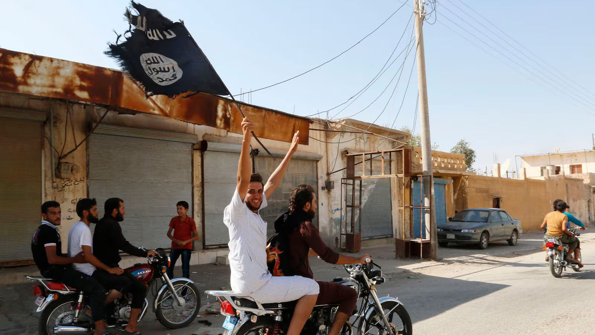 A man rides on the back of a motorcycle waving a black ISIS flag through a street in northern Syria as bystanders watch.