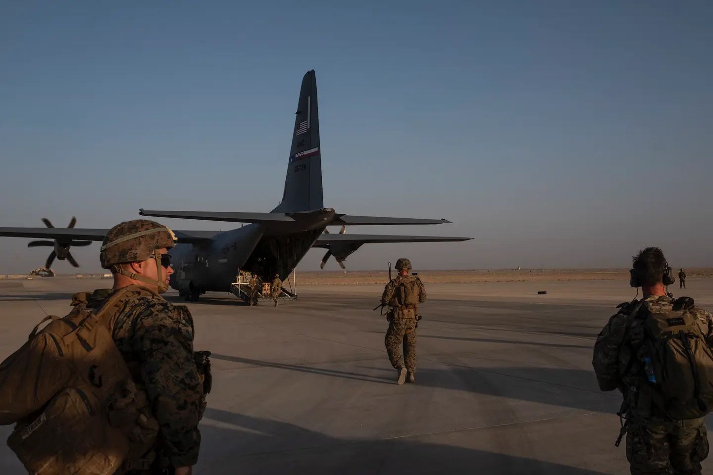 U.S. Marines carrying packs and equipment walk toward a U.S. Air Force C-130 Hercules transport aircraft on a runway in Afghanistan during military operations, with the desert landscape stretching into the distance.