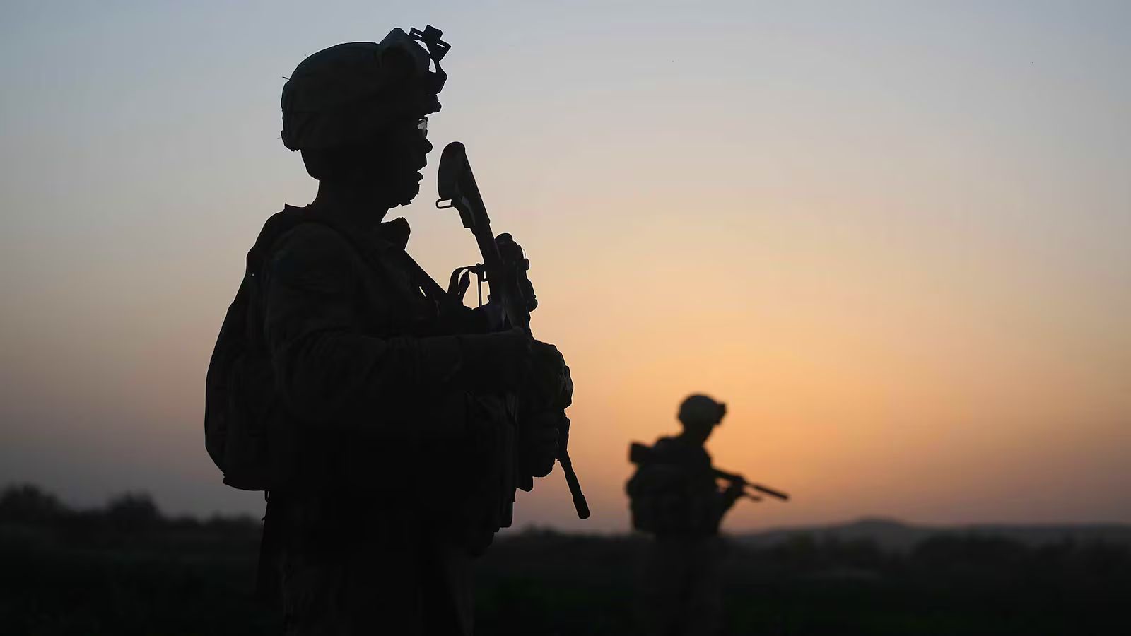 Silhouettes of two U.S. soldiers on patrol at sunset, with one service member standing in the foreground holding a rifle while another moves through the distance across an open landscape.