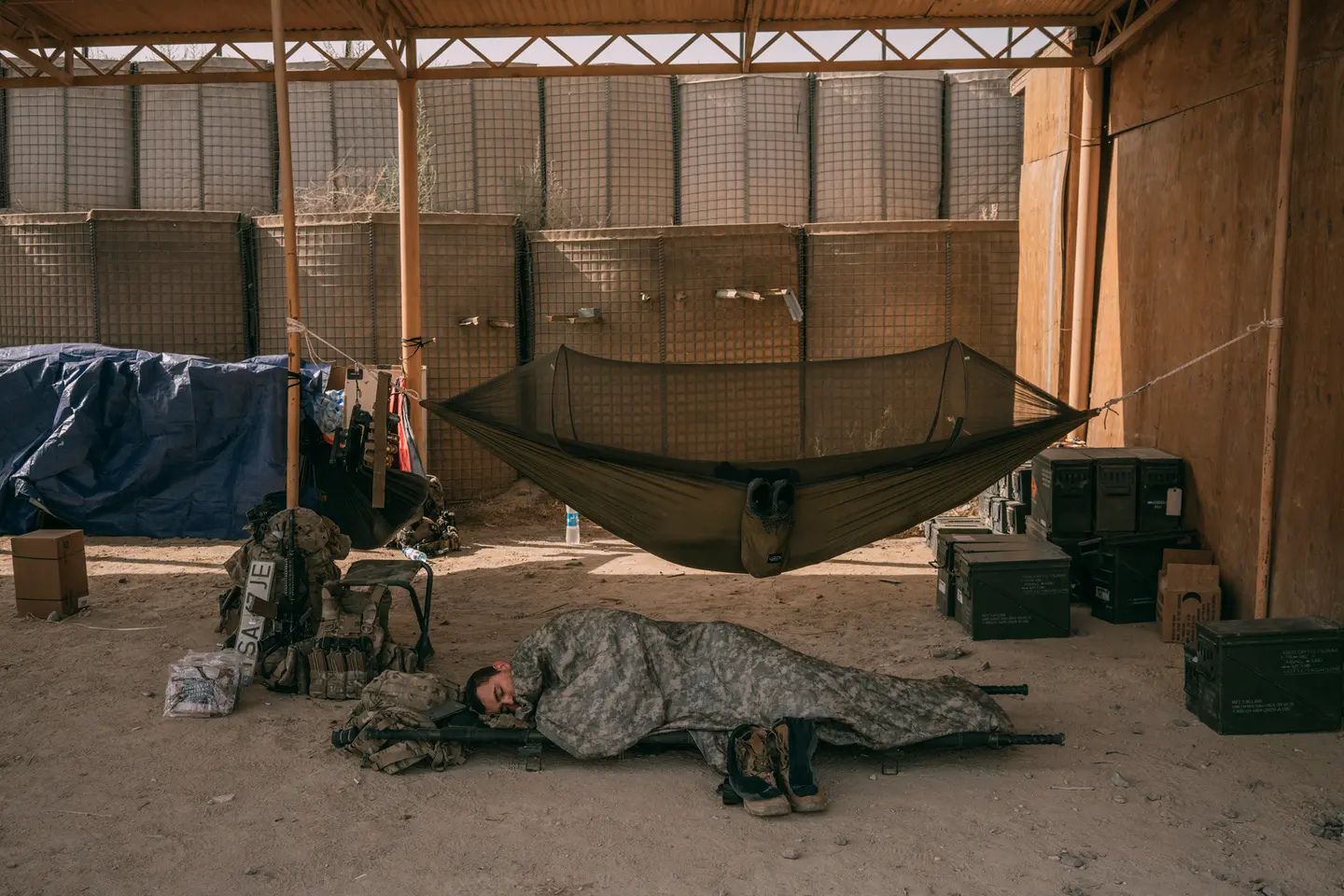 U.S. service member sleeps on a field cot beneath a camouflage blanket inside a fortified combat outpost in Afghanistan.