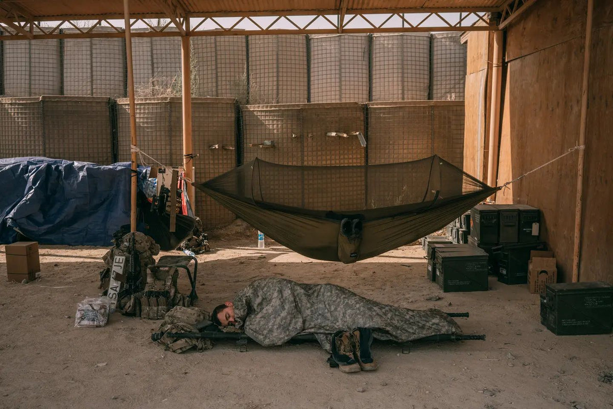 U.S. service member sleeps on a field cot beneath a camouflage blanket inside a fortified combat outpost in Afghanistan.