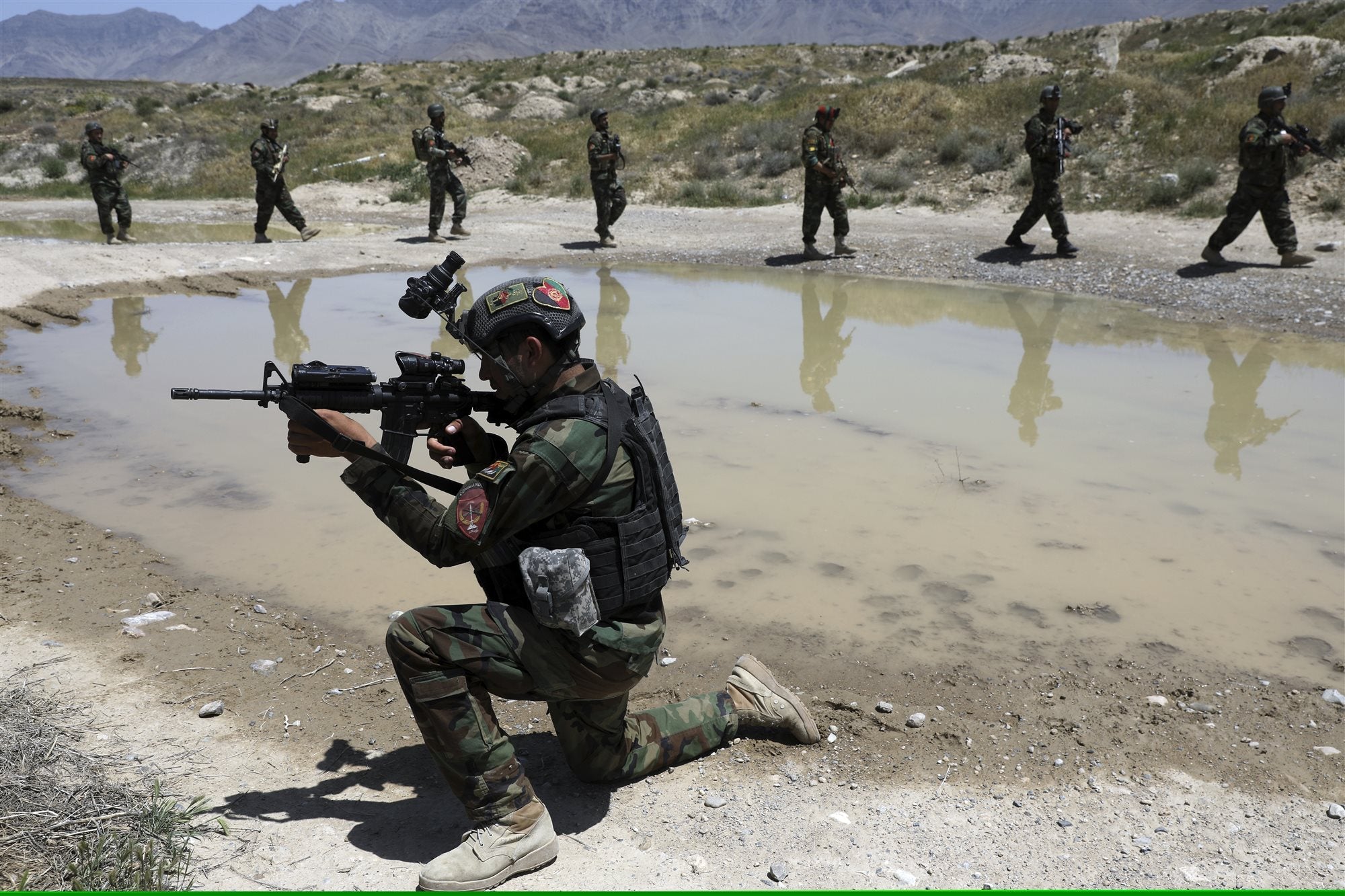 An Afghan National Army commando kneels with a rifle raised during a training exercise, with soldiers patrolling in the background.